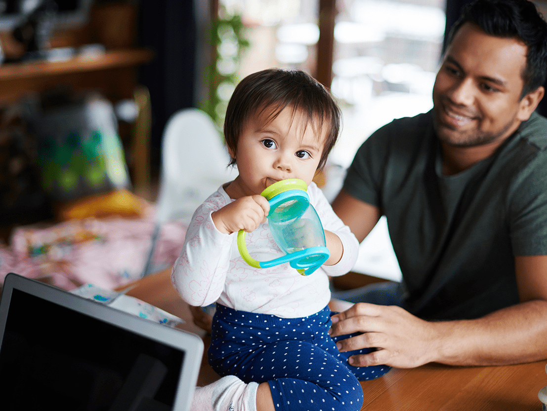 baby sitting on table drinking water with father in sitting next to her